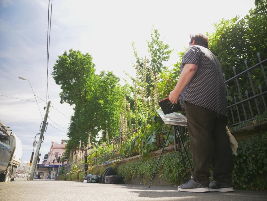 A man standing on a footpath with easel, painting the scene up the street, lots of greenery around.