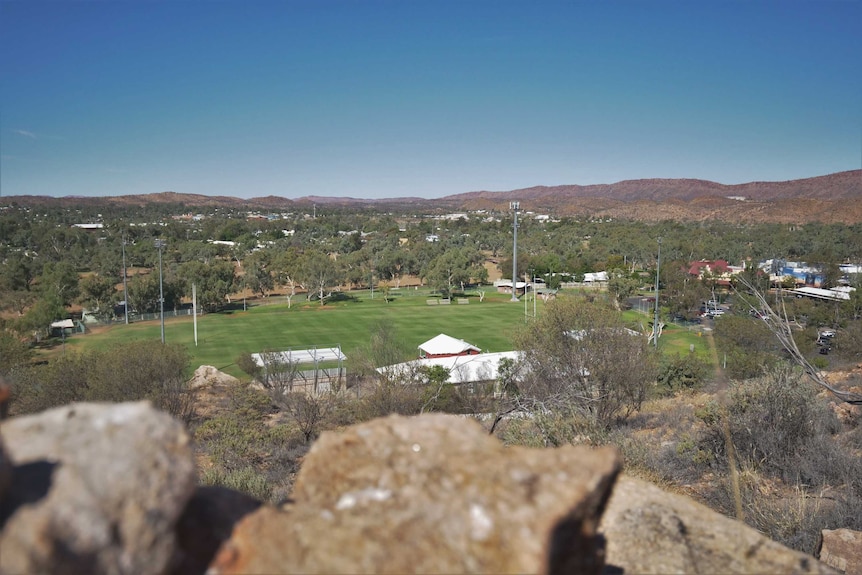 A view from the top of a hill down to a green oval in a town.