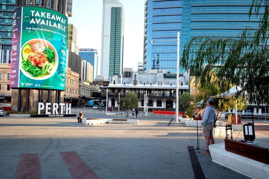 A man wearing a hat stands in front of a microphone singing in Yagan Square in the Perth CBD.