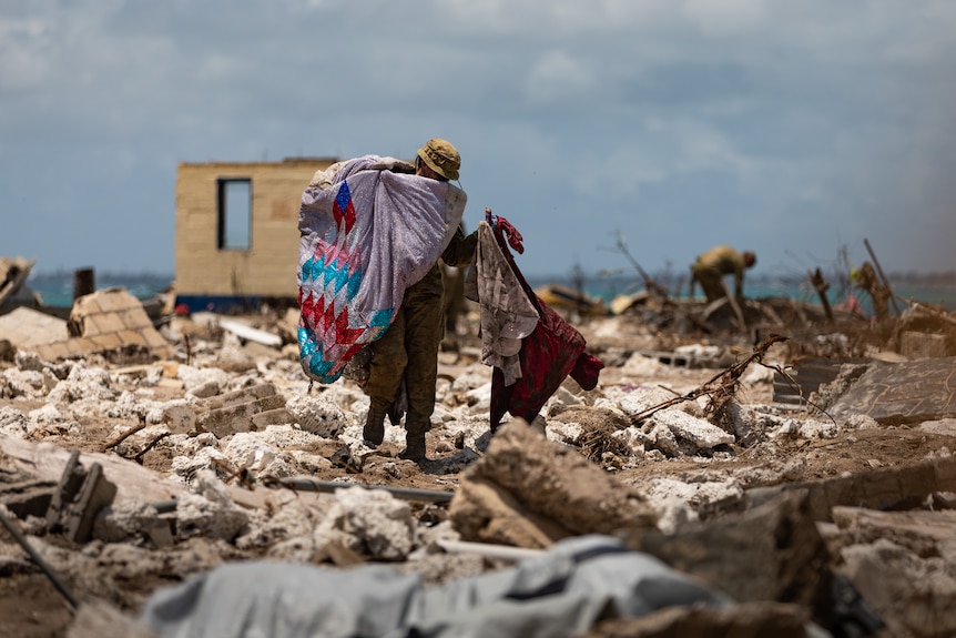 A soldier walks through an area of complete wreckage carrying debris.
