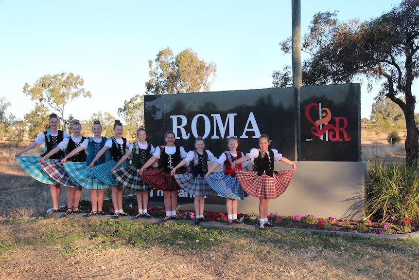 Nine girls, dressed in highland dancing costumes, stand in front of the Roma town sign.