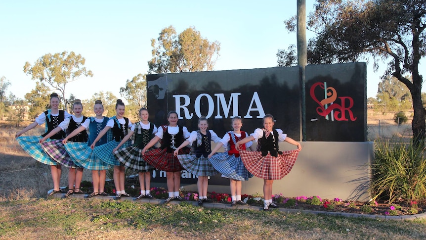 Nine girls, dressed in highland dancing costumes, stand in front of the Roma town sign.