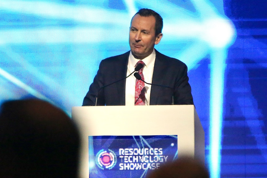 WA Premier Mark McGowan at a podium on a stage in front of a blue electronic screen delivering a speech and wearing a suit.
