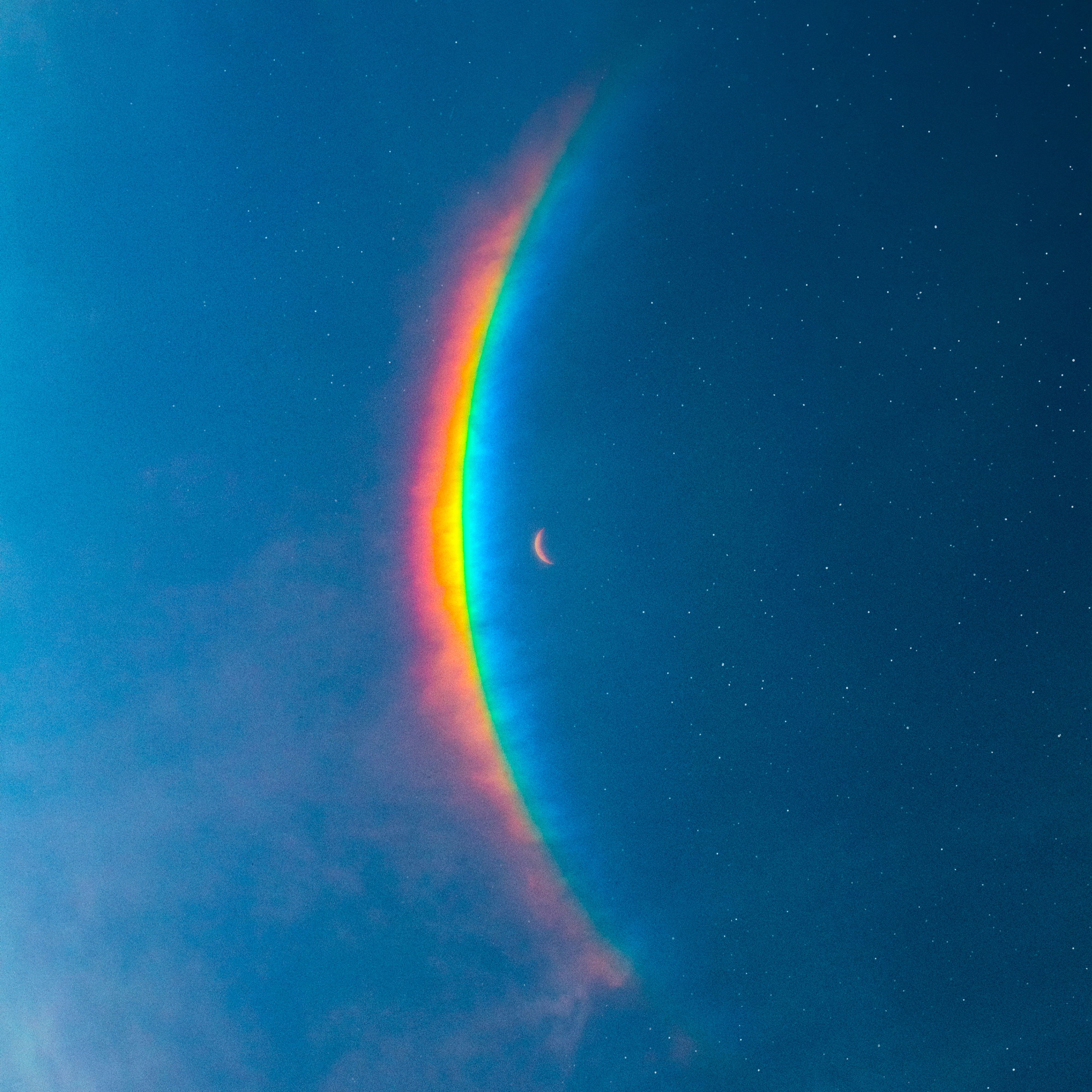 A shot of the atmosphere where a crescent rainbow has formed in the ozone, a crescent moon can be seen in the distance