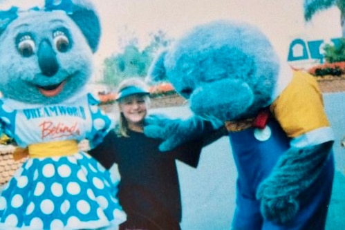 A blurred photo of a child at Dreamworld, smiling as she is flanked by two costume koalas in blue polka dot dress and overalls.