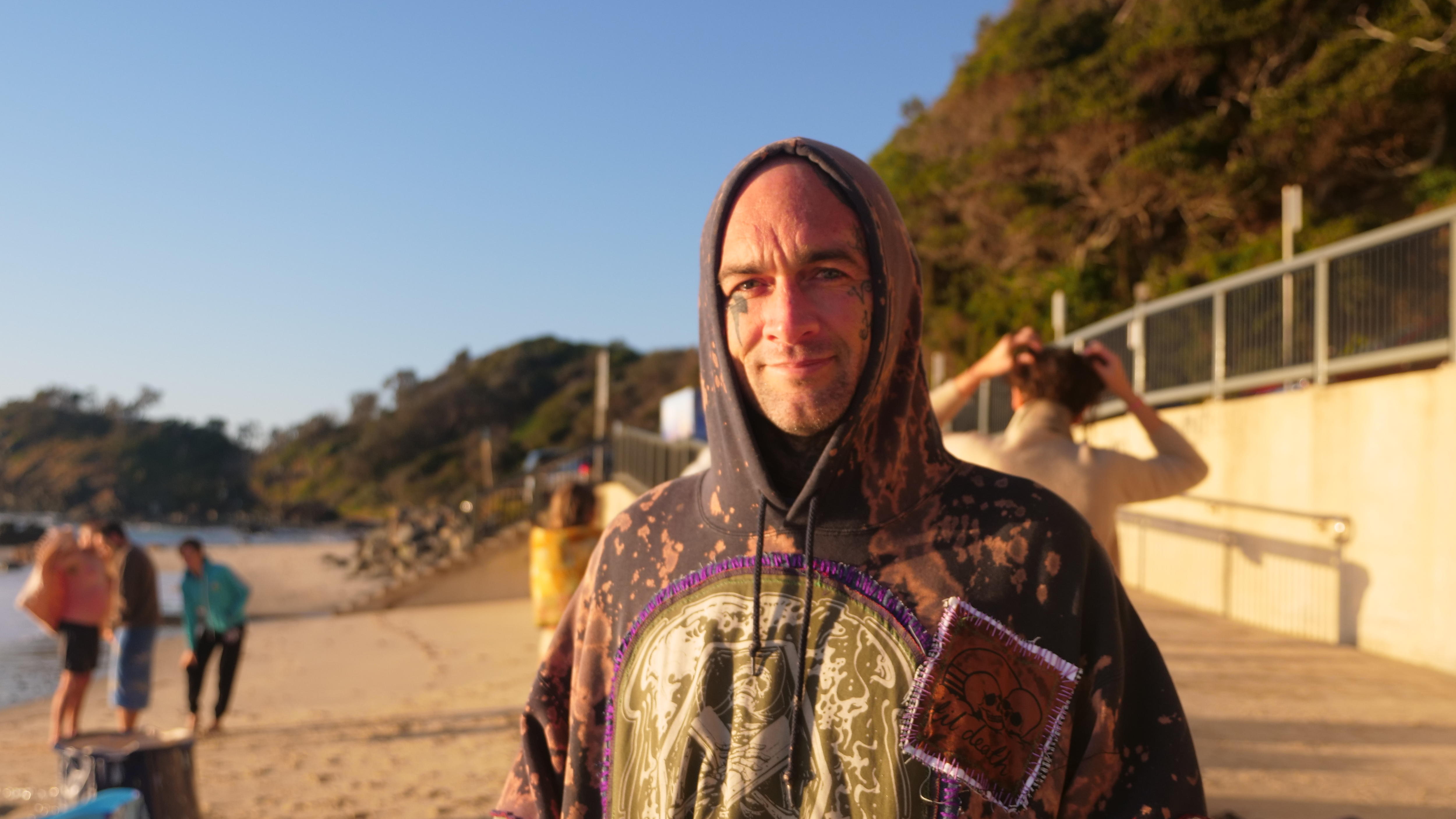 A man with tattoos on his face stands on a beach in a hoodie