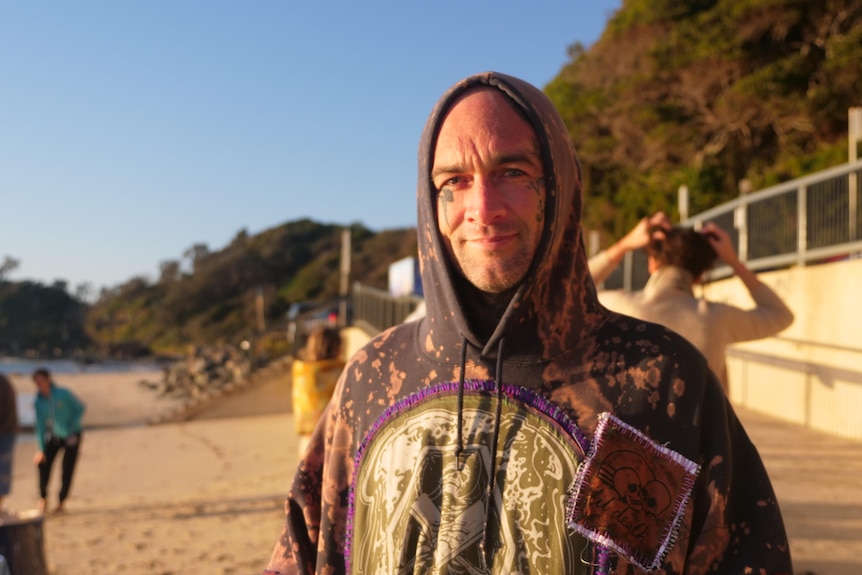 A man with tattoos on his face stands on a beach in a hoodie