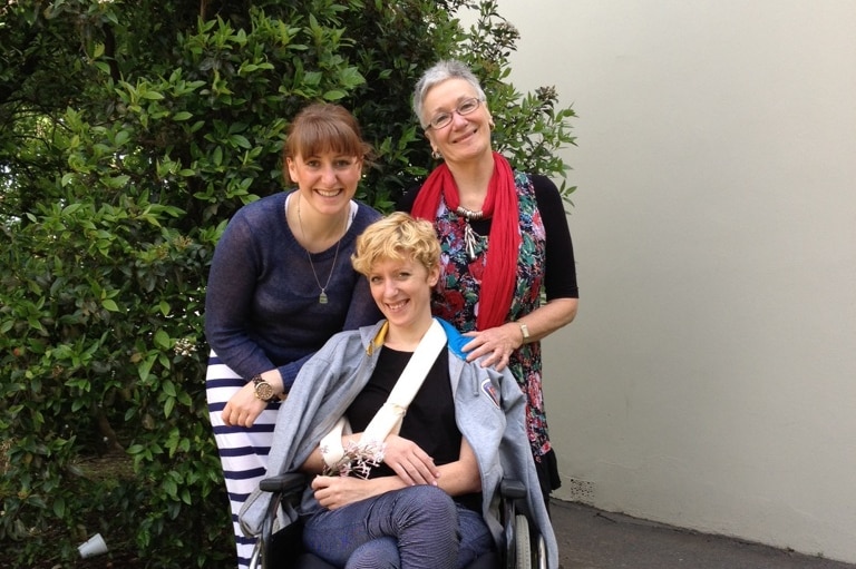 Jemma Burns sits in a wheelchair smiling with her mother Kerri Greening and sister Firlie Greening standing behind her.