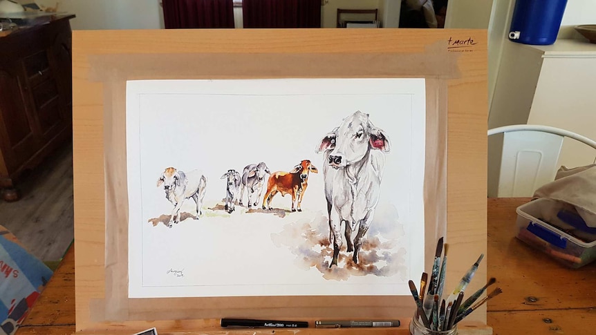 A wooden easel rests on a table with a watercolour picture of Beryl the Brahman and four other cattle standing behind her in mud