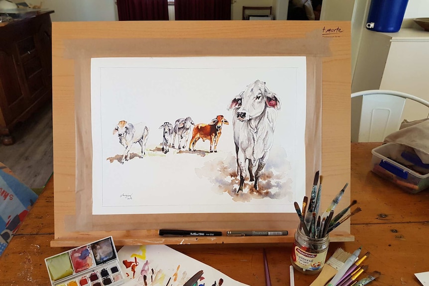 A wooden easel rests on a table with a watercolour picture of Beryl the Brahman and four other cattle standing behind her in mud