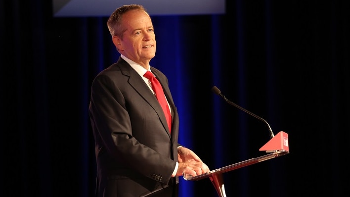 A man in a dark suit and red tie speaks from behind a lectern on a stage in front of drapes and political banners