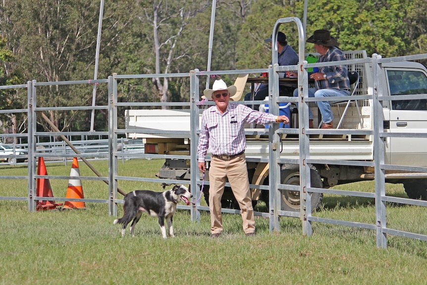 A man in a check shirt and hat opens gate to leave show ring with his black and white dog.