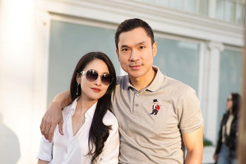 A close up of a man wearing a button up shirt with his arm around a woman wearing sunglasses.