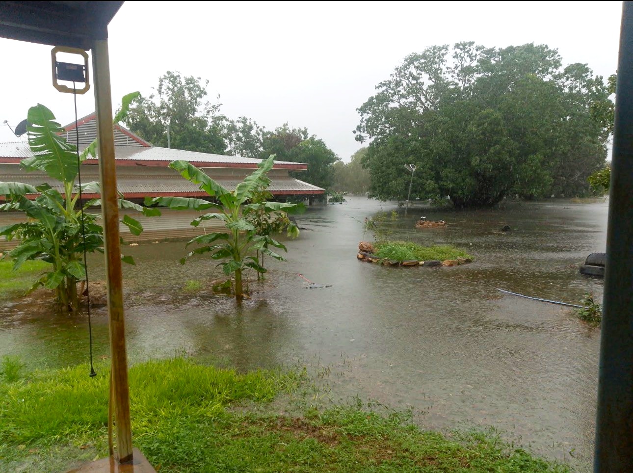House in back left of image under water, halfway up its front porch, flood waters, small green trees in foreground.