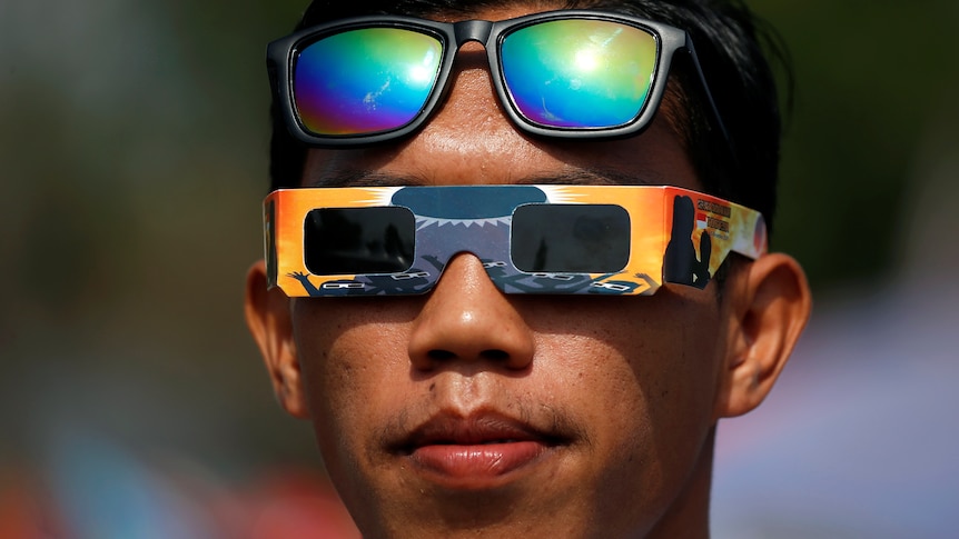 A man wears special solar eclipse glasses on his eyes, with his sunglasses resting up higher on his head. 