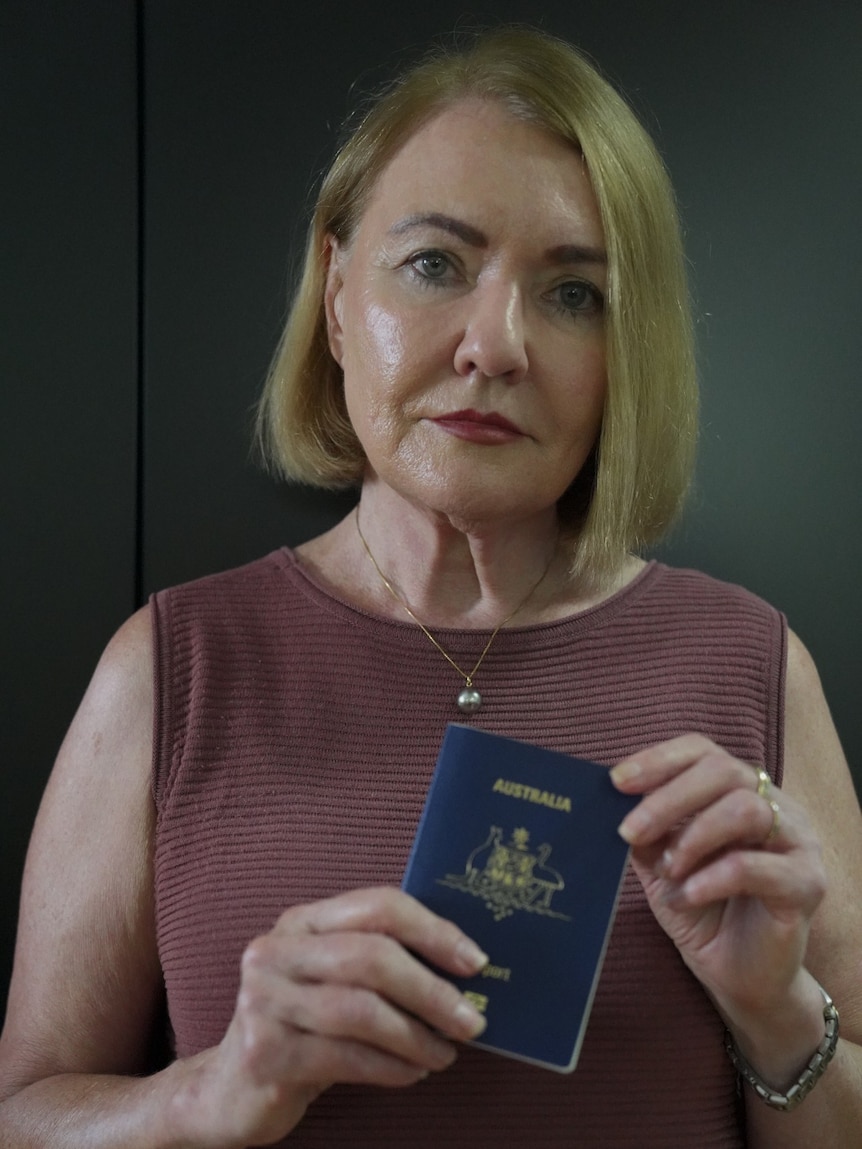 A woman with mid-length blonde hair holds her passport and looks at the camera.