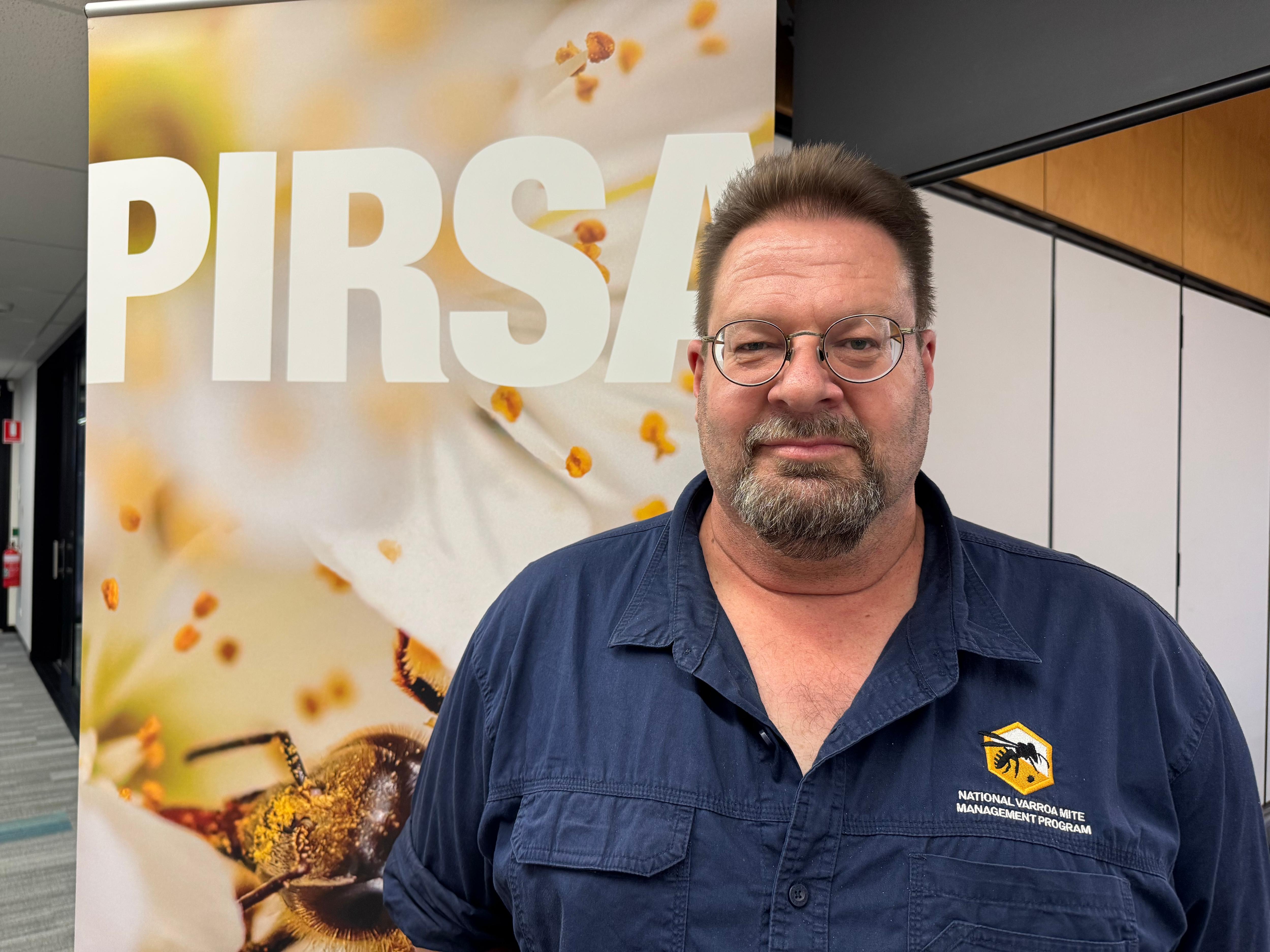 A middle-aged, bearded man with dark hair wears a dark, branded work shirt while standing in front of a sign that says "PIRSA".