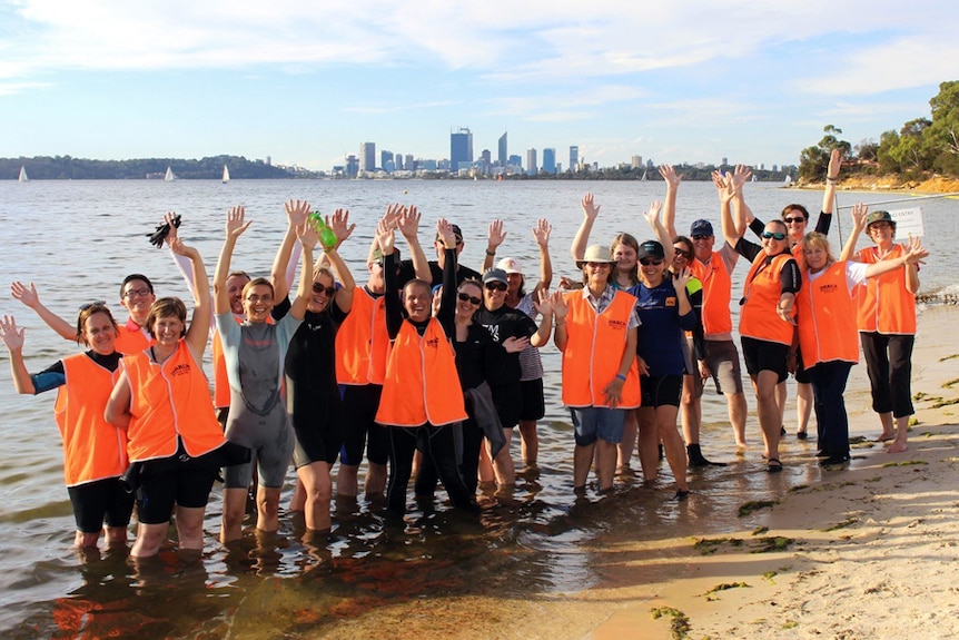 A grouop of people in hi-viz vest on the shore of the Swan River with Perth in the background