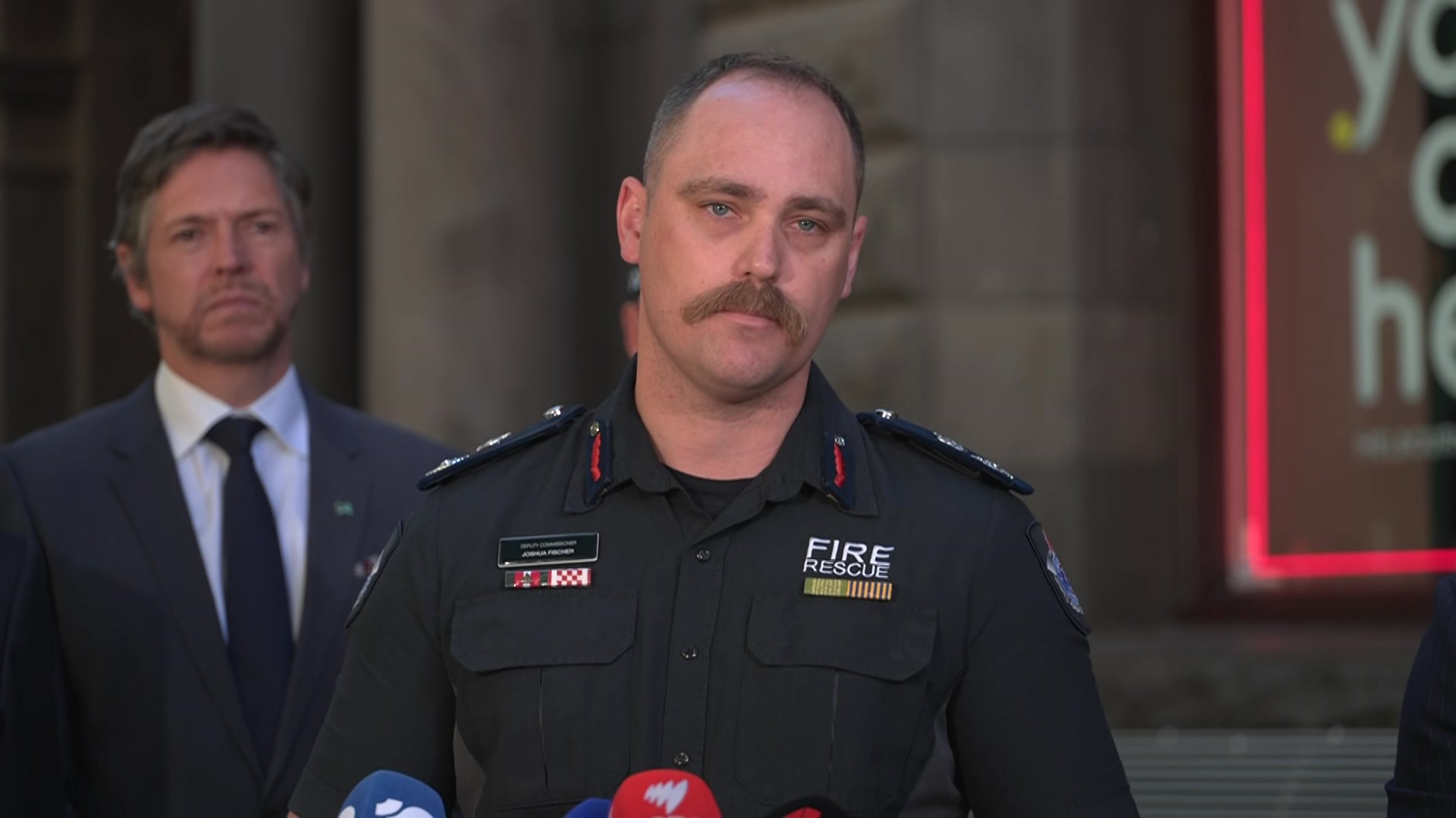 Um homem de cabelos castanhos e bigode, vestindo uma blusa azul com distintivos e uma escrita que diz "resgate de incêndio" Ele está localizado atrás dos microfones.