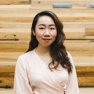 A young Asian woman in a light pink shirt standing in front of a wooden wall.