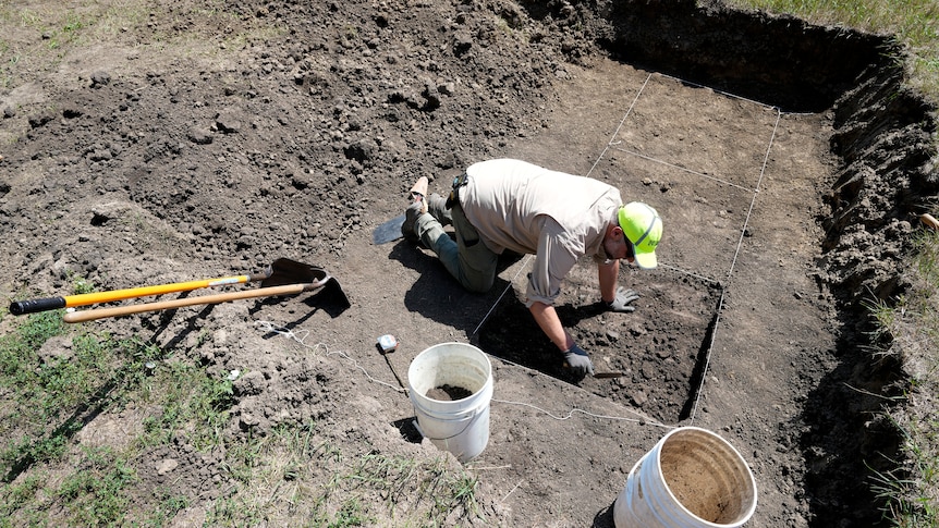 A white man in dull clothing and a bright yellow cap kneels on the ground as he works in a square hole at a dig site.