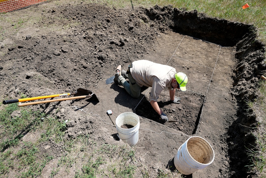 A white man in dull clothing and a bright yellow cap kneels on the ground as he works in a square hole at a dig site.