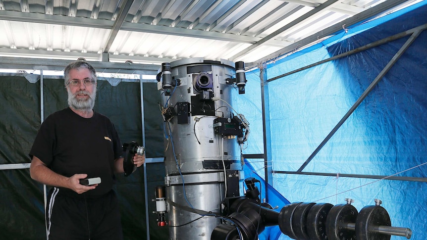 A man wearing all black and aviator sunglasses stands next to a silver telescope which is taller