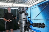 A man wearing all black and aviator sunglasses stands next to a silver telescope which is taller