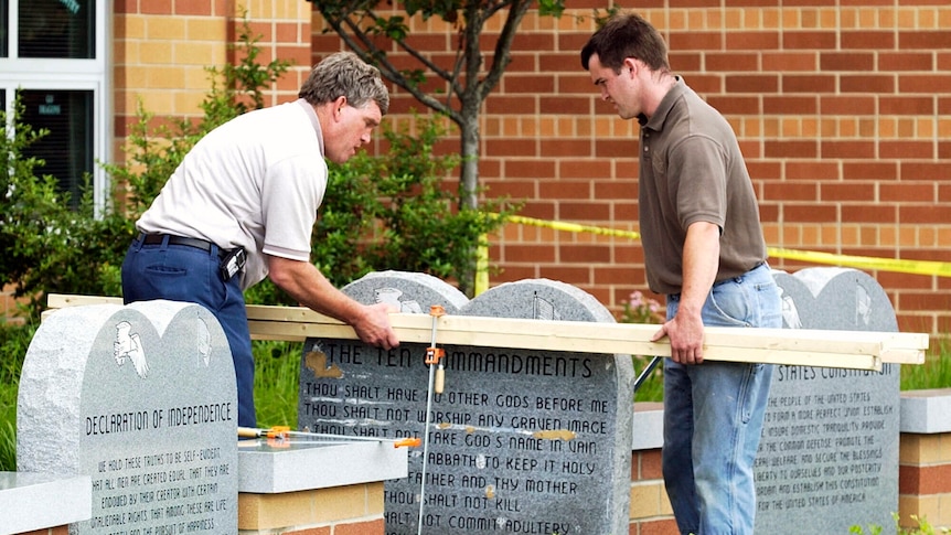 Two men measure a stone tablet bearing the Ten Commandments outside a brick building.