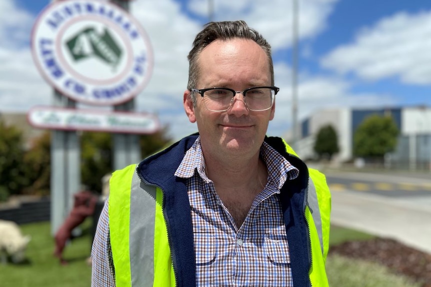 A man in black-rimmed glasses, a checked shirt and yellow high-vis vest stands outside a factory.