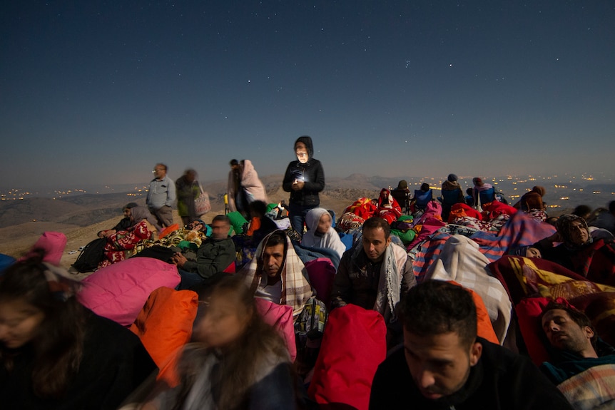 A crowd of people in colourful clothing and warm blankets huddle together on a rocky mountain top as stars twinkle in the sky.