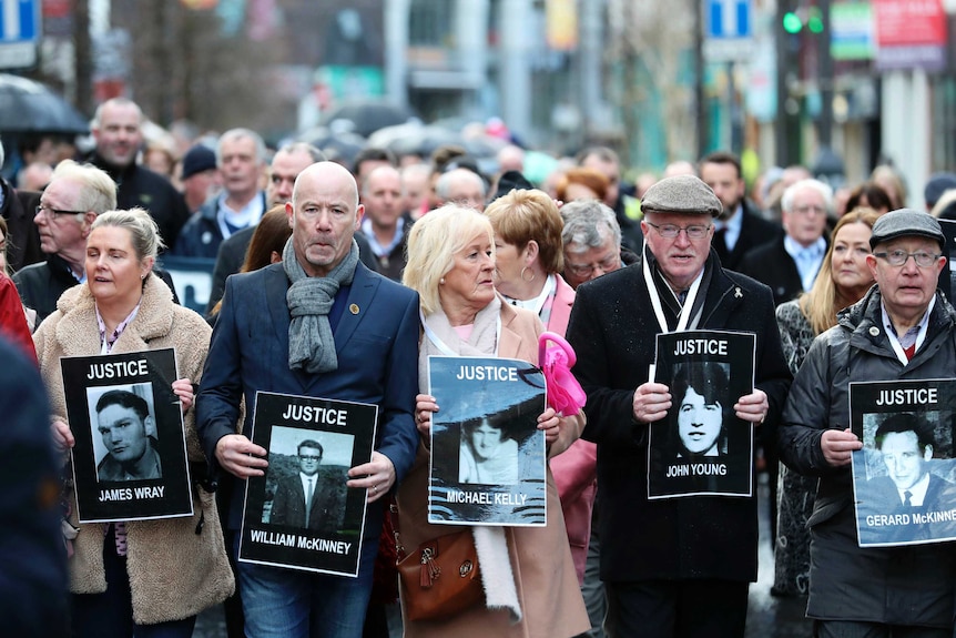 People march in the street carrying large black-and-white photos of their family members with their names and the word 'justice'