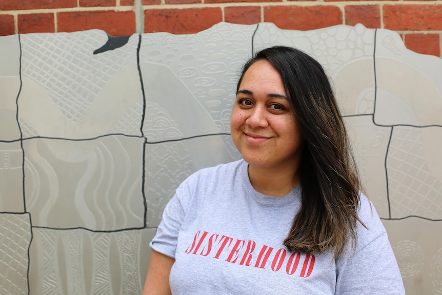 A woman with brown skin and hair, wearing a grey t-shirt that says 'sisterhood' stands in front of a brick wall, smiling. 