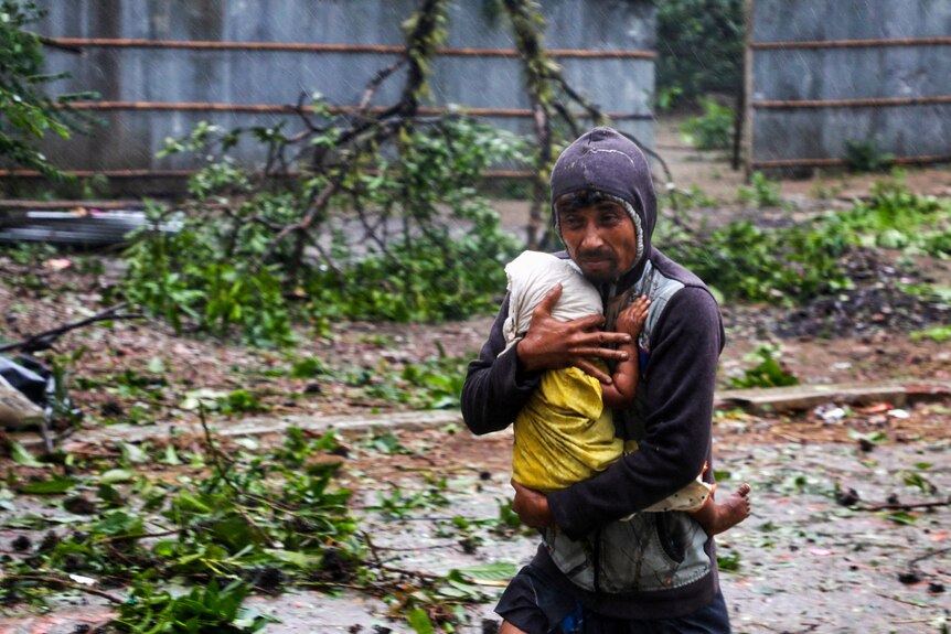 A man cradles a child and walks while trees and debris lie in the background during a storm.