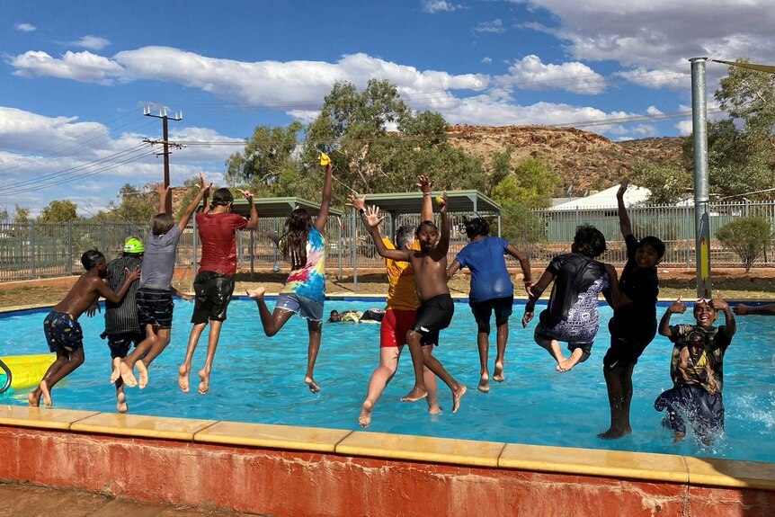 A group of about a dozen children smile as they jump into a pool with their arms up