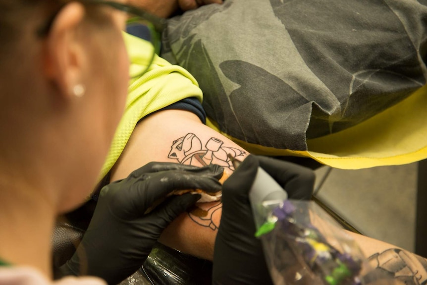 Ash chain begins to add colour to the outline of a Pokemon character she is tattooing on the inside of a man's bicep.