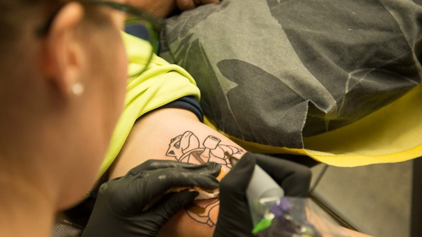 Ash chain begins to add colour to the outline of a Pokemon character she is tattooing on the inside of a man's bicep.