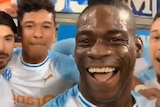 Mario Balotelli smiles at the camera with his teammates behind him and fans in the background