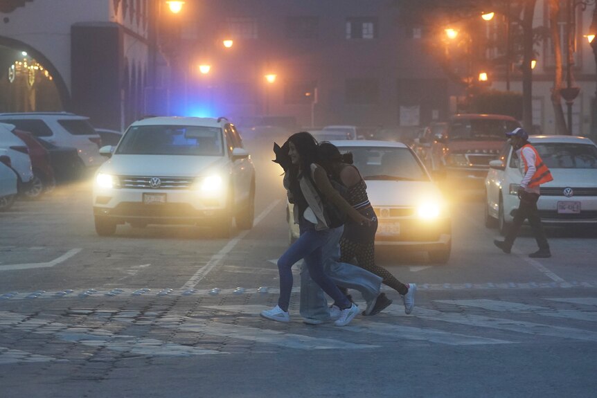 Women walk along a pedestrian crossing past cars with headlights on, with a haze in the air.