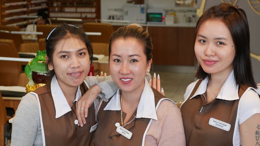 Three women working at a nail salon pose for the camera in front of the business.