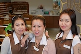 Three women working at a nail salon pose for the camera in front of the business.