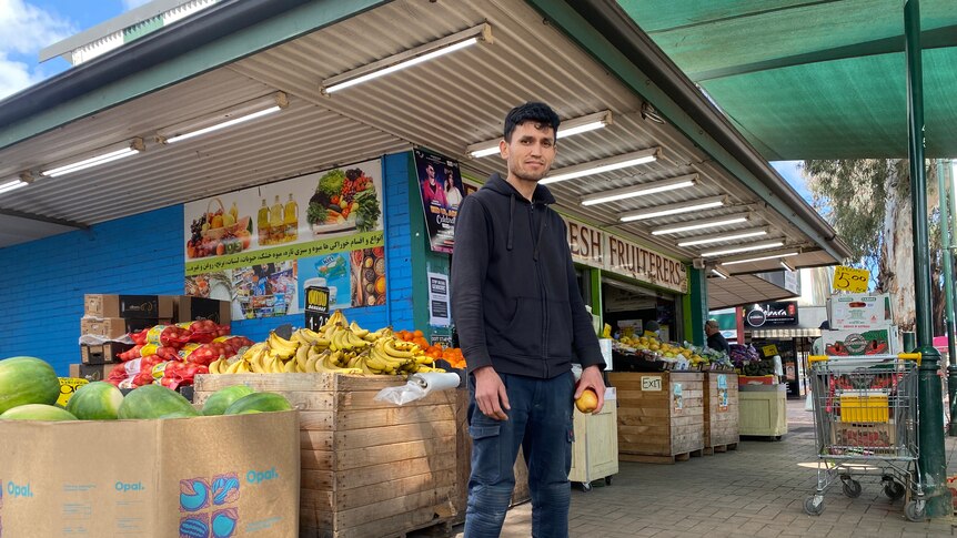 A man stands in front of a fruit and vegetable shop, holding an apple in his hand