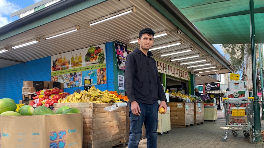 A man stands in front of a fruit and vegetable shop, holding an apple in his hand