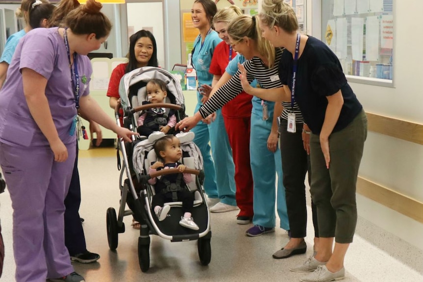 Nurses line up and wave to twin girls in a pram being pushed by their mother in a hospital corridor