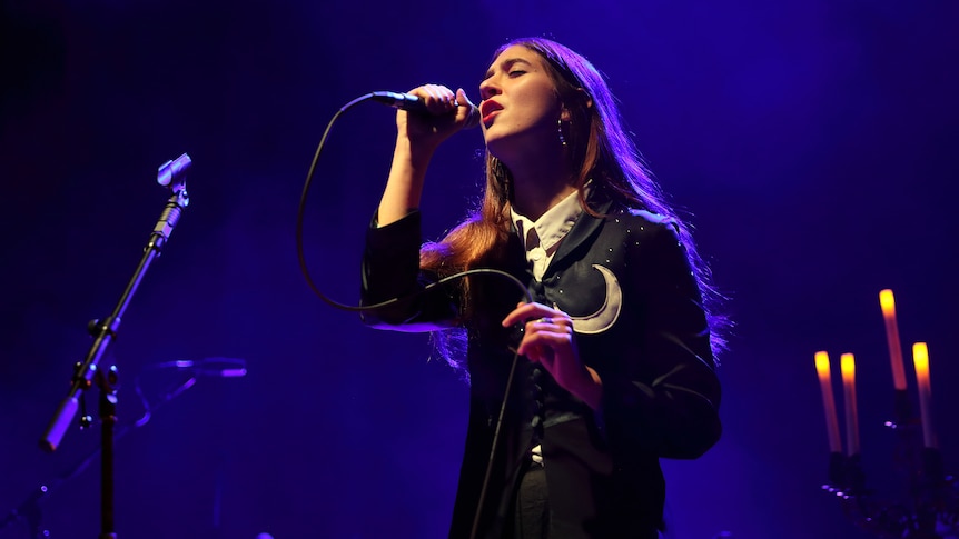 Natalie Mering stands holding a microphone. She has long straight hair and a black jacket