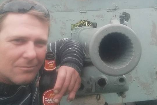 A man with sunglasses atop his head poses with a beer next to an historic cannon.
