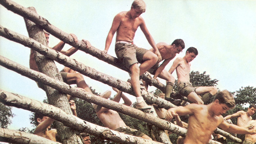 A bunch of teenage boys climb over a wooden obstacle.