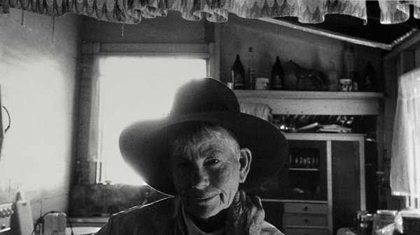 Black and white photograph of an elderly woman in a cowboy hat, sitting in a rural kitchen.