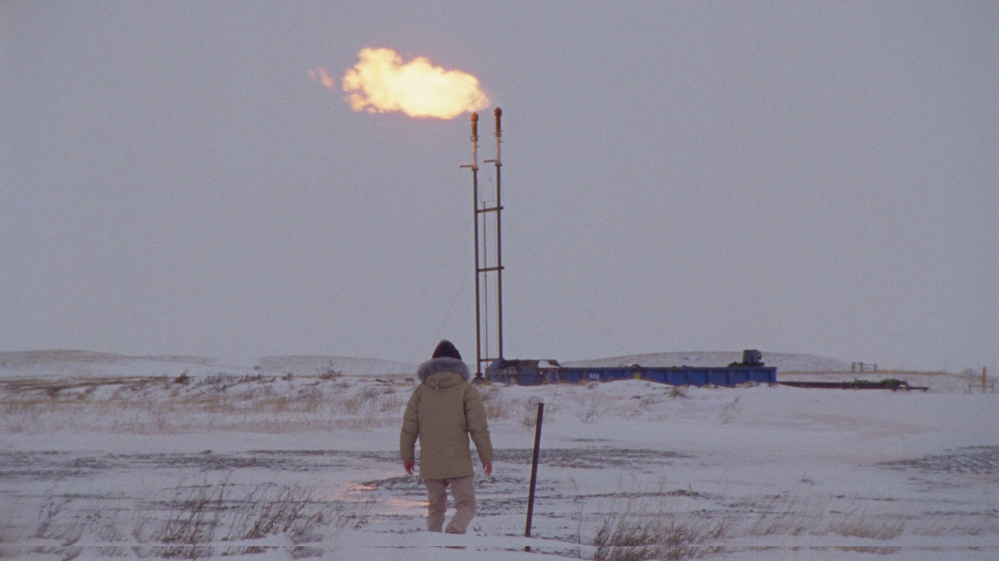 A person dressed in a taupe weather-beater jacket and beanie stands in a barren snowy landscape with a burning oil pipeline.