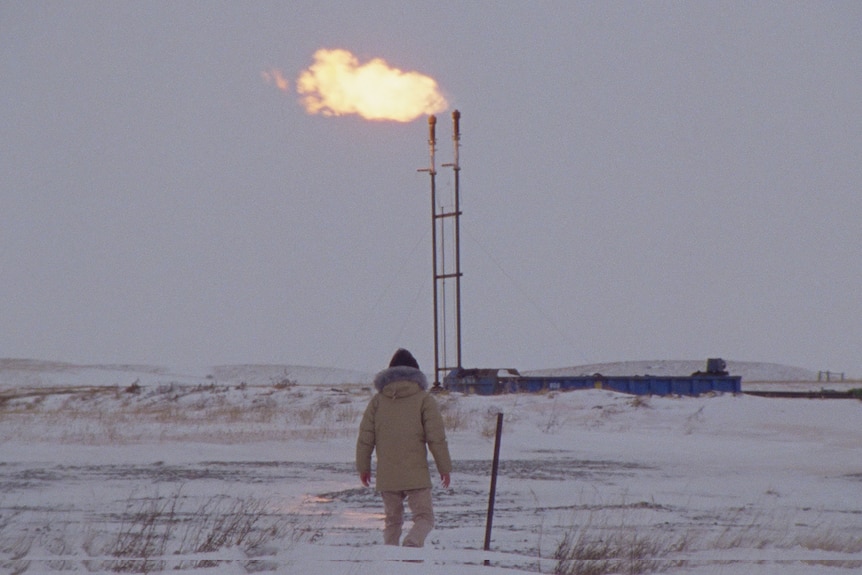 A person dressed in a taupe weather-beater jacket and beanie stands in a barren snowy landscape with a burning oil pipeline.